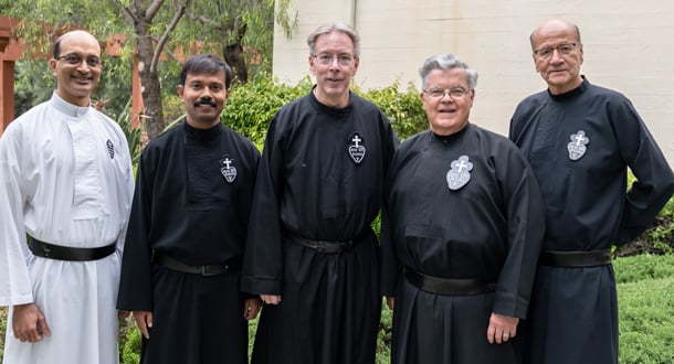 Five Passionist priests stand side by side outdoors in front of greenery and a light-colored building. Four are wearing traditional black Passionist habits with the emblem on the chest, and one on the far left is wearing a white habit with a black belt. They are smiling and facing the camera in a relaxed, friendly pose.