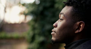 A young man with short curly hair and a trimmed beard is shown in profile, gazing thoughtfully into the distance. The background is softly blurred with greenery and light, suggesting an outdoor setting.