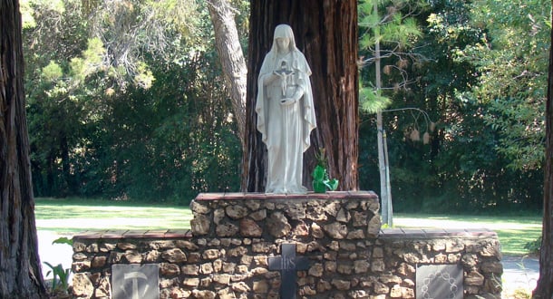 Outdoor statue of the Virgin Mary holding a crucifix, placed on a stone pedestal with a cross, surrounded by tall trees and greenery in the background.