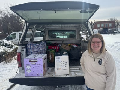 A smiling woman stands beside an open truck bed filled with donated supplies, including diapers, a box fan, and bags of various items. The scene takes place on a snowy day, with snow covering the ground and rooftops in the background.