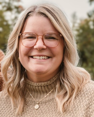 Portrait of a smiling woman with long blonde hair, wearing glasses and a tan knit sweater with layered necklaces, standing outdoors with greenery in the background.