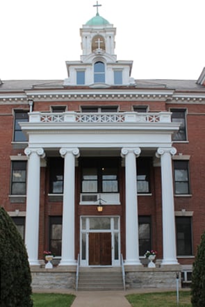 Front view of a red-brick monastery building with tall white columns, a central wooden door, and a cupola topped with a cross.