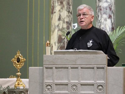 Fr. Jack Conley, CP, in a Passionist habit at a marble pulpit inside a church. Behind him are tall marble columns, a microphone, and a golden monstrance placed on a side altar.