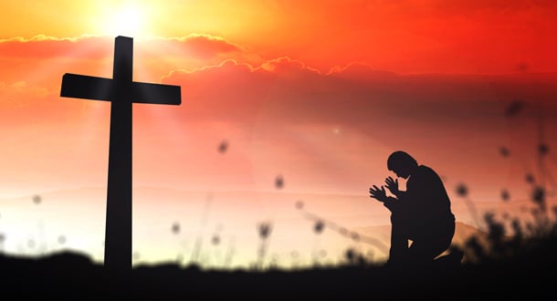 Silhouette of a person kneeling in prayer beside a large cross, set against a vivid orange and red sunset sky.