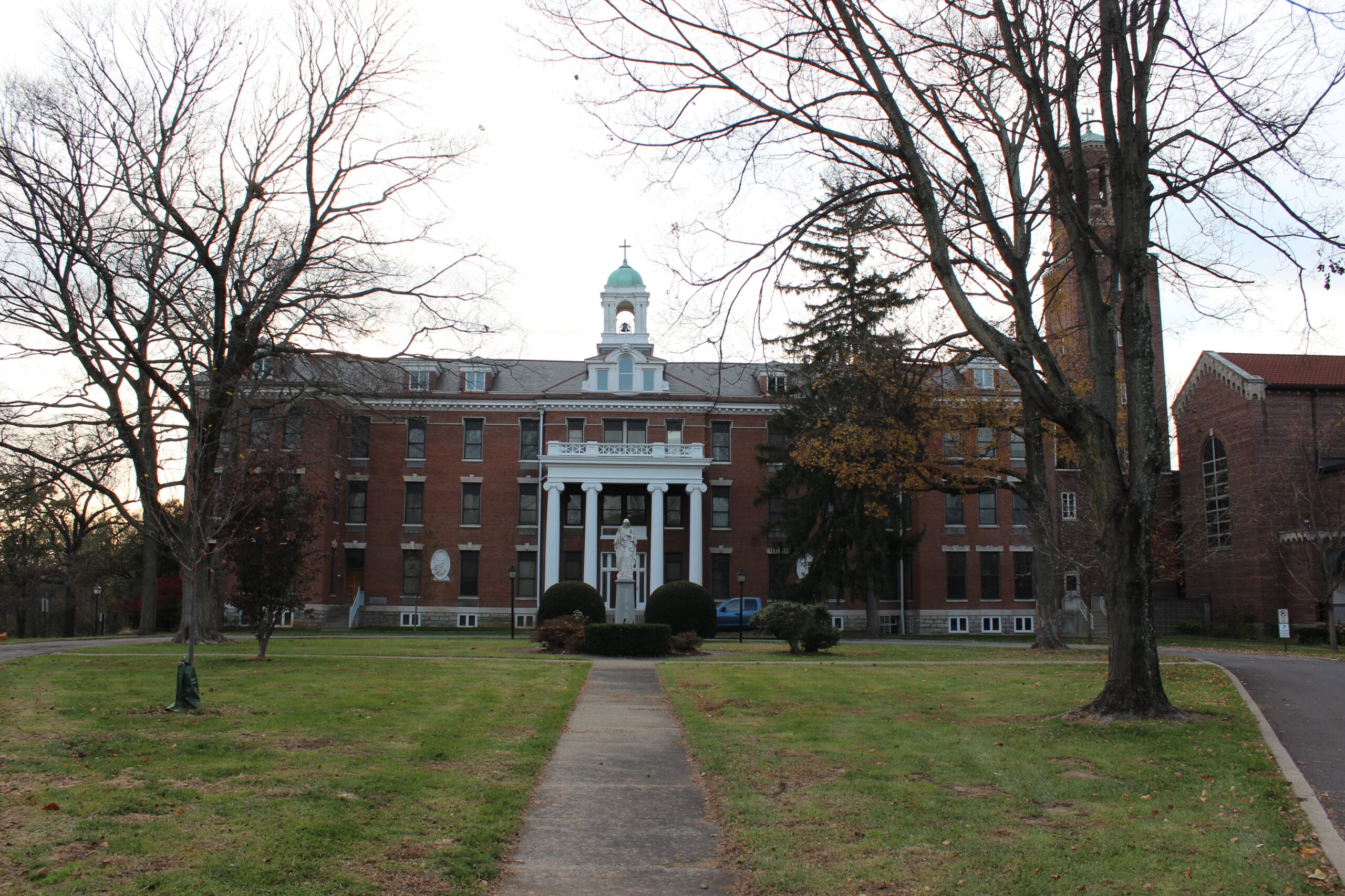 A large red-brick monastery with white columns and a green-domed bell tower stands at the end of a tree-lined path, flanked by leafless trees and manicured lawns.