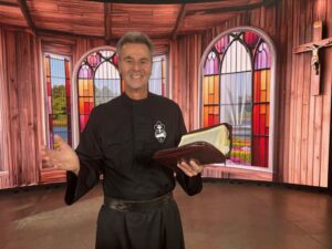 Smiling Fr. Cedric Pisegna, a Passionist Priest, in a studio with stained glass and a crucifix