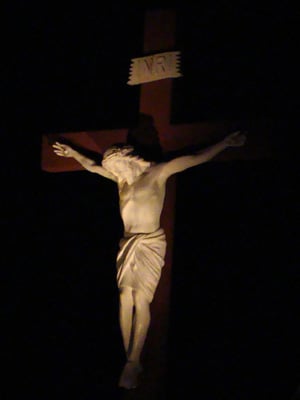 A spotlighted crucifix depicting Jesus Christ crucified on a wooden cross, set against a dark background. A sign marked "INRI" is affixed above Christ's head.