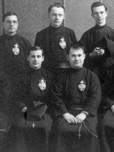 A black-and-white historical photograph of six Passionist priests wearing traditional habits featuring the emblem of the Passionist order. The men are posed formally, some seated and some standing.