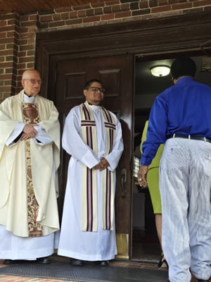 Two priests stand at the entrance of a church, one in a cream chasuble and the other in a white alb with a striped stole, greeting two parishioners as they enter. The setting is warm and welcoming.