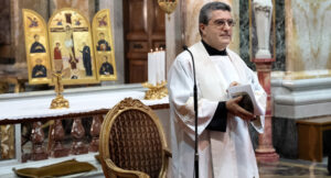 A priest in white vestments holds a book while standing near the altar, with candles and a gold religious icon in the background.