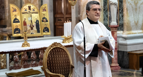 A priest in white vestments holds a book while standing near the altar, with candles and a gold religious icon in the background.