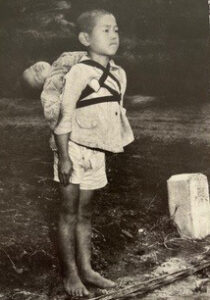 Historic black-and-white photograph of a solemn young boy standing barefoot, carrying his deceased baby brother on his back after the atomic bombing of Nagasaki in 1945.