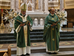 Two priests in green liturgical vestments stand before the altar inside a church, reading from books during a service.