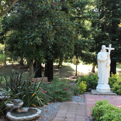 A peaceful garden with a white statue of a saint holding a cross, surrounded by greenery, flowers, a stone fountain, and tall trees in the background.