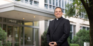 A smiling Catholic priest in black clerical attire stands in front of the entrance to the Catholic Theological Union building, surrounded by greenery.