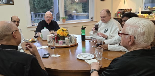 Passionist priests gather around a round table sharing a meal and conversation in a warm, communal setting.