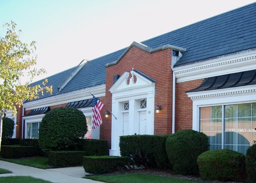 A red-brick office building with white trim and a gabled entrance displays an American flag near the front door, surrounded by neatly trimmed hedges and a sidewalk.