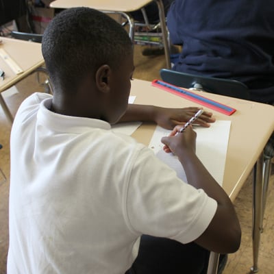 Young student writing at a desk in a classroom, wearing a white polo shirt and focused on his paper.