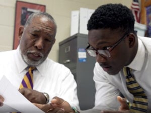 Teacher Mentoring Student. An older man and a younger man sit side-by-side at a desk, reviewing a document together.