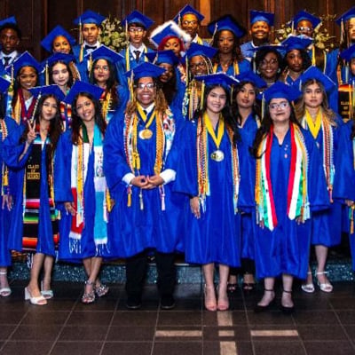 Group of high school graduates in blue caps and gowns posing for a celebratory class photo indoors.