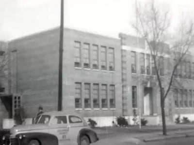 Black and white photo of a mid-20th-century school building with a vintage car parked in front, labeled “Ambulance 85.”