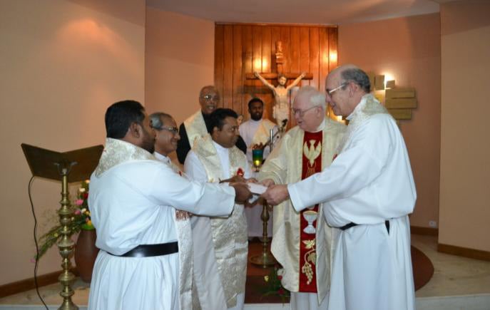 A group of Catholic priests, dressed in white and cream liturgical vestments, stand together during a ceremony in a chapel. Two priests are exchanging documents or vows while others look on. A crucifix mounted on a wooden panel is visible in the background, emphasizing the sacred setting.