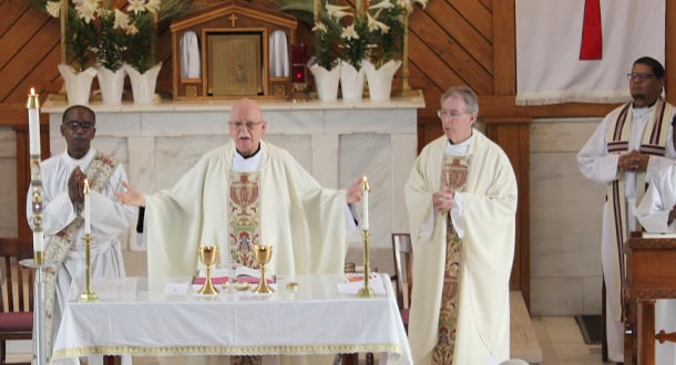 Four clergy members in white vestments stand at the altar during Mass, with candles, flowers, and chalices arranged on the altar.
