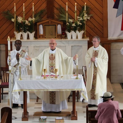 Two priests and a deacon in white vestments stand at the altar during Mass at Holy Family Parish, with candles, flowers, and liturgical vessels arranged on the altar, and a few parishioners visible in the foreground.