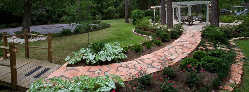 A landscaped garden with a stone walkway leading past flower beds and greenery to a pergola with seating, next to a small wooden footbridge.
