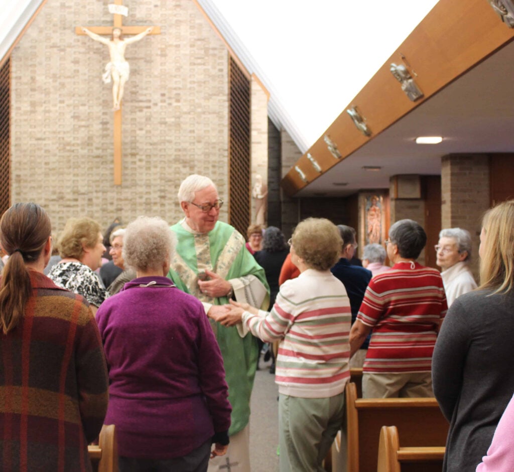 A priest in green vestments stands in the center aisle of a church, warmly greeting and shaking hands with parishioners after Mass. The church is filled with older adults standing in pews, facing the priest. A large crucifix hangs on the brick wall at the front of the sanctuary. The atmosphere is warm and communal.