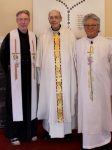 Three priests stand together smiling, dressed in liturgical vestments—two in white albs and one in a black habit with a white stole—inside a church.
