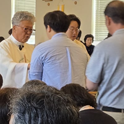 A priest in a white alb distributes communion to parishioners during a Catholic Mass, with a full congregation in the pews of a church.