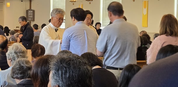 A priest in a white alb distributes communion to parishioners during a Catholic Mass, with a full congregation in the pews of a church.