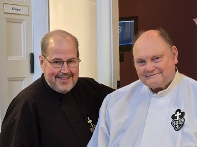 Two Passionist priests, one in a black habit and the other in white, smile while standing near a chapel door.