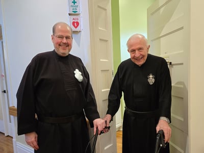 Two Passionist priests in black habits stand together in a hallway, smiling, one using a walker.