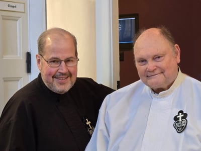 Two Passionist priests stand together smiling, one in a black habit and the other in a white habit with the Passionist emblem, inside a chapel entryway.