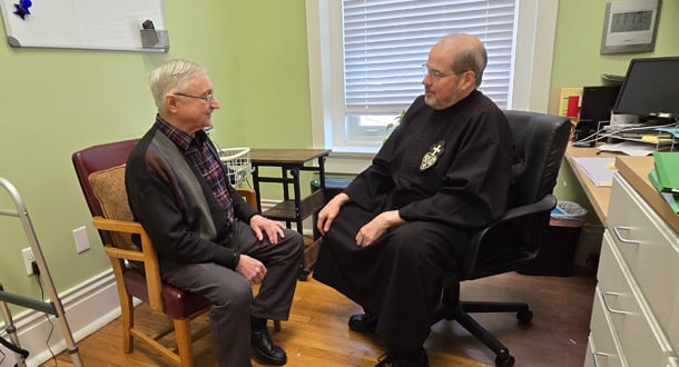 A Passionist brother in black habit sits in an office speaking with an older man seated across from him.