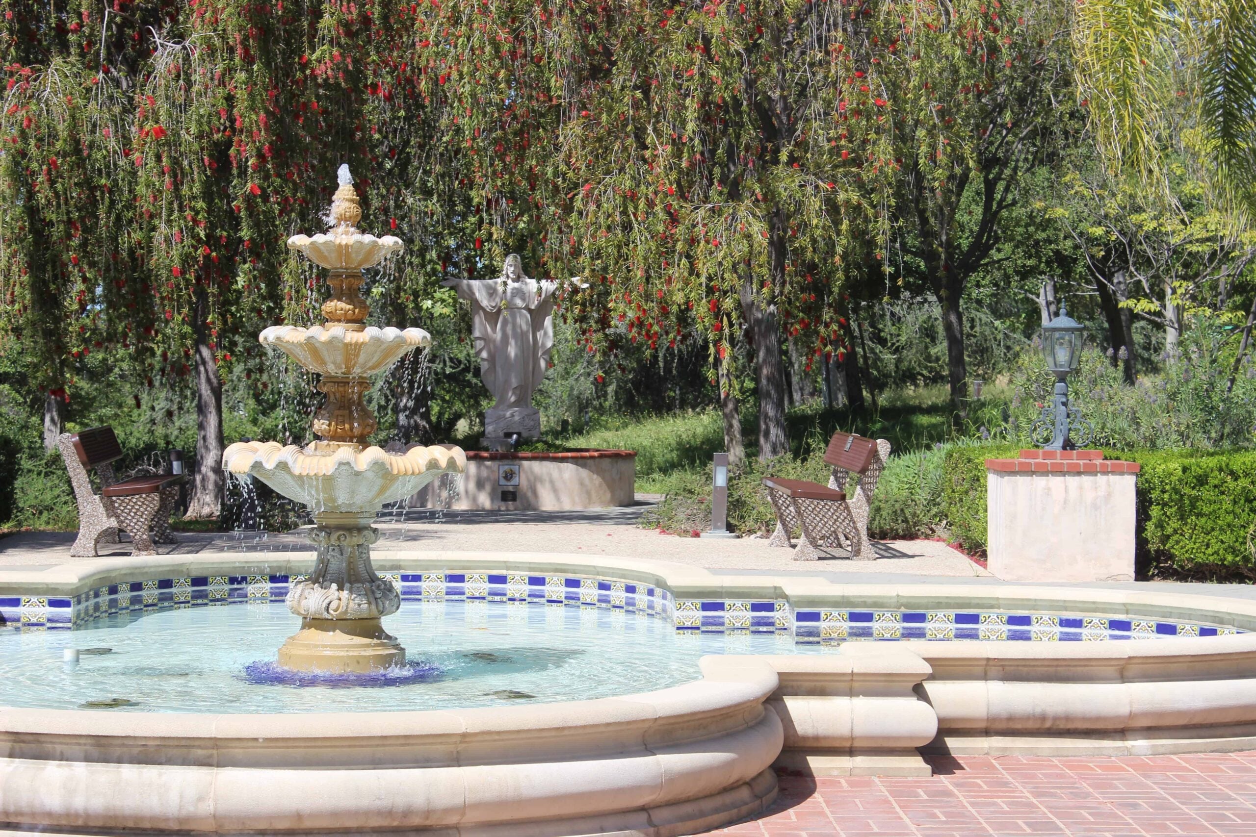 A three-tiered fountain with water flowing into a tiled basin, set in a courtyard with benches, flowering trees, and a statue of Jesus with outstretched arms in the background.