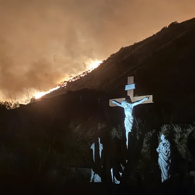 A crucifix and statues illuminated in the foreground as a wildfire burns on the hillside behind them at night.