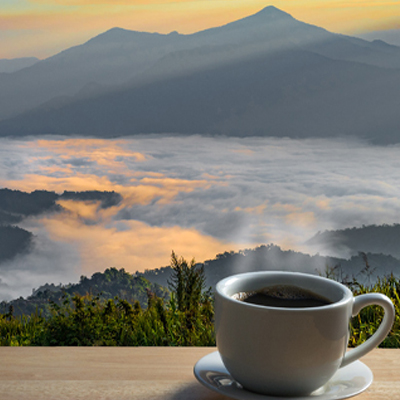 A steaming cup of coffee on a table overlooking a valley filled with morning mist, with mountains silhouetted against a colorful sunrise.