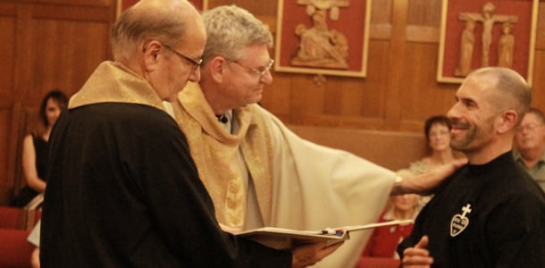 A Passionist priest places a hand on a young man in habit during a profession ceremony inside a chapel.