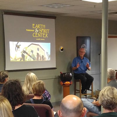 A speaker addresses a seated audience during a presentation at the Earth & Spirit Center, with a projection screen and whiteboard visible in a brick-walled room.