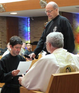 A young man in religious habit makes his profession before a seated priest during a ceremony, while another priest holds a microphone.
