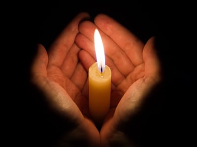 Close-up of two cupped hands gently holding a lit candle in the dark, the flame casting a warm glow.