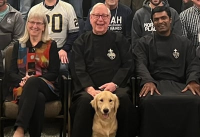 A woman and two Passionist priests sit smiling with a golden retriever in front of them, surrounded by a group.
