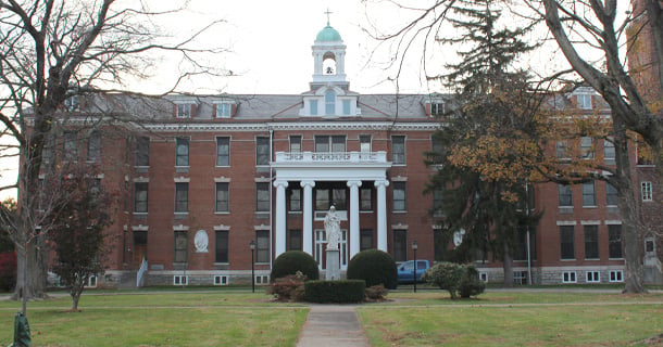 A front-facing view of Sacred Heart Monastery, a large red-brick building with white columns and a central statue of the Sacred Heart of Jesus. The structure features a green-domed cupola and is surrounded by bare trees and neatly trimmed bushes on a spacious lawn.
