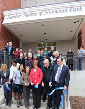 A group of community leaders, clergy, and officials gather for a ribbon-cutting ceremony on the steps of the Senior Suites of Norwood Park, celebrating its grand opening.