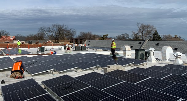 Workers install rows of solar panels on a large flat rooftop under a partly cloudy sky.