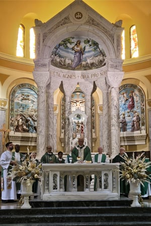 Catholic priests celebrate Mass at the ornate marble altar of St. Agnes Church, surrounded by intricate mosaics, religious artwork, and golden light streaming through stained glass windows.