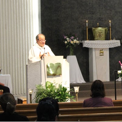 Priest delivering a homily at the ambo in a church, with congregation seated and altar in the background.